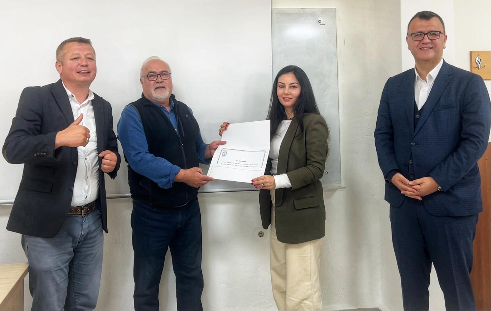This photograph, taken in a classroom, shows a middle-aged man handing a document to a woman in the center and two other men watching the documents with smiles. The woman is serious and polite as she hands over the document. The other man standing next to them has his hands clasped in front of him and is wearing a suit.